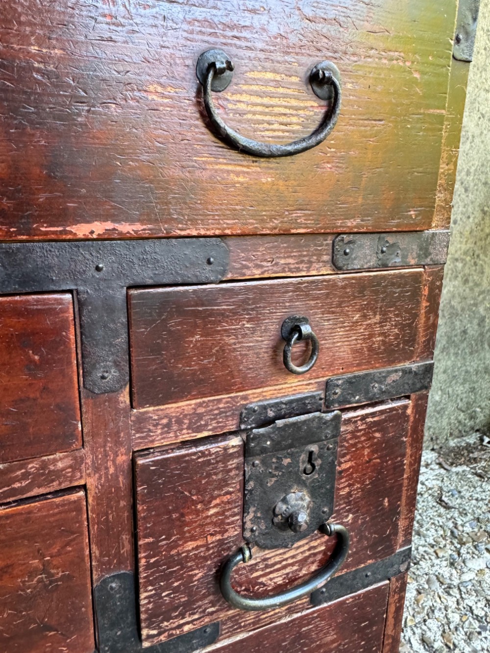 A reddish brown lacquered Japanese wooden tansu chest featuring iron corner plates, circular lock fittings, side carrying handles, and multiple graduated drawers. View VII
