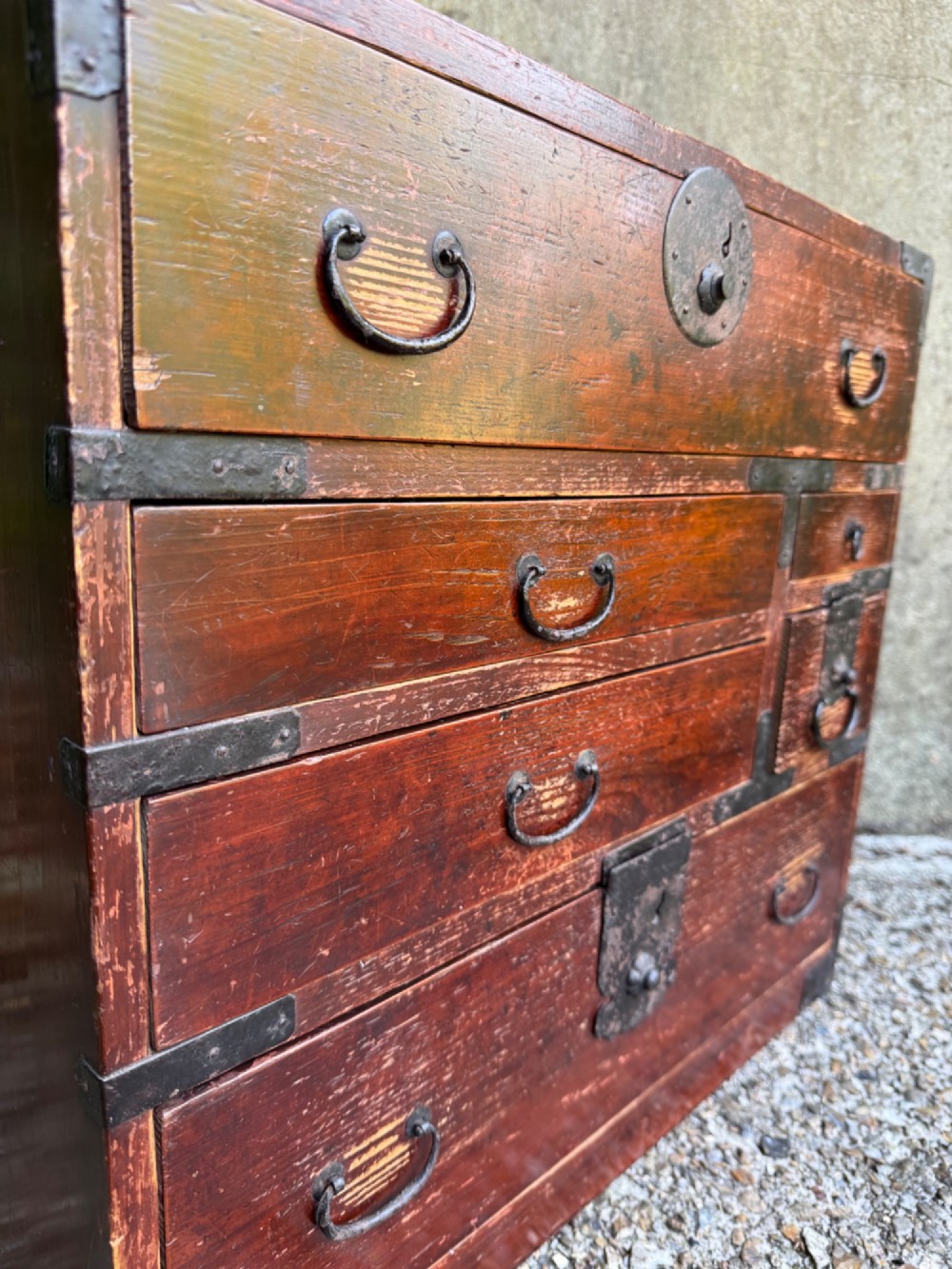 A reddish brown lacquered Japanese wooden tansu chest featuring iron corner plates, circular lock fittings, side carrying handles, and multiple graduated drawers. View X