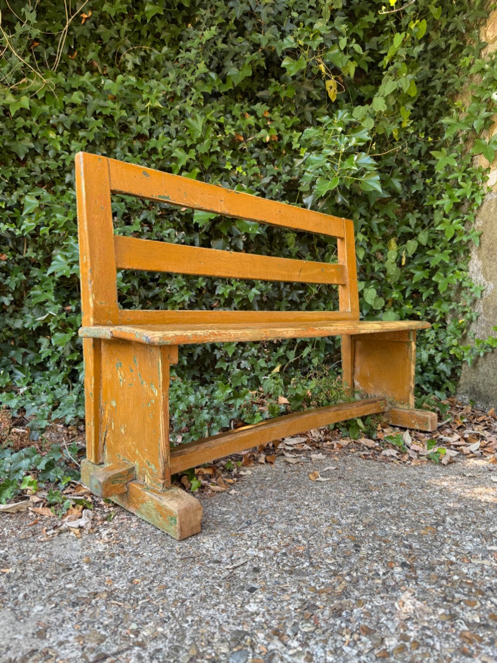 An antique rustic pine bench with a horizontal slat back, distressed mustard yellow painted finish, and solid end supports joined by a lower stretcher.
