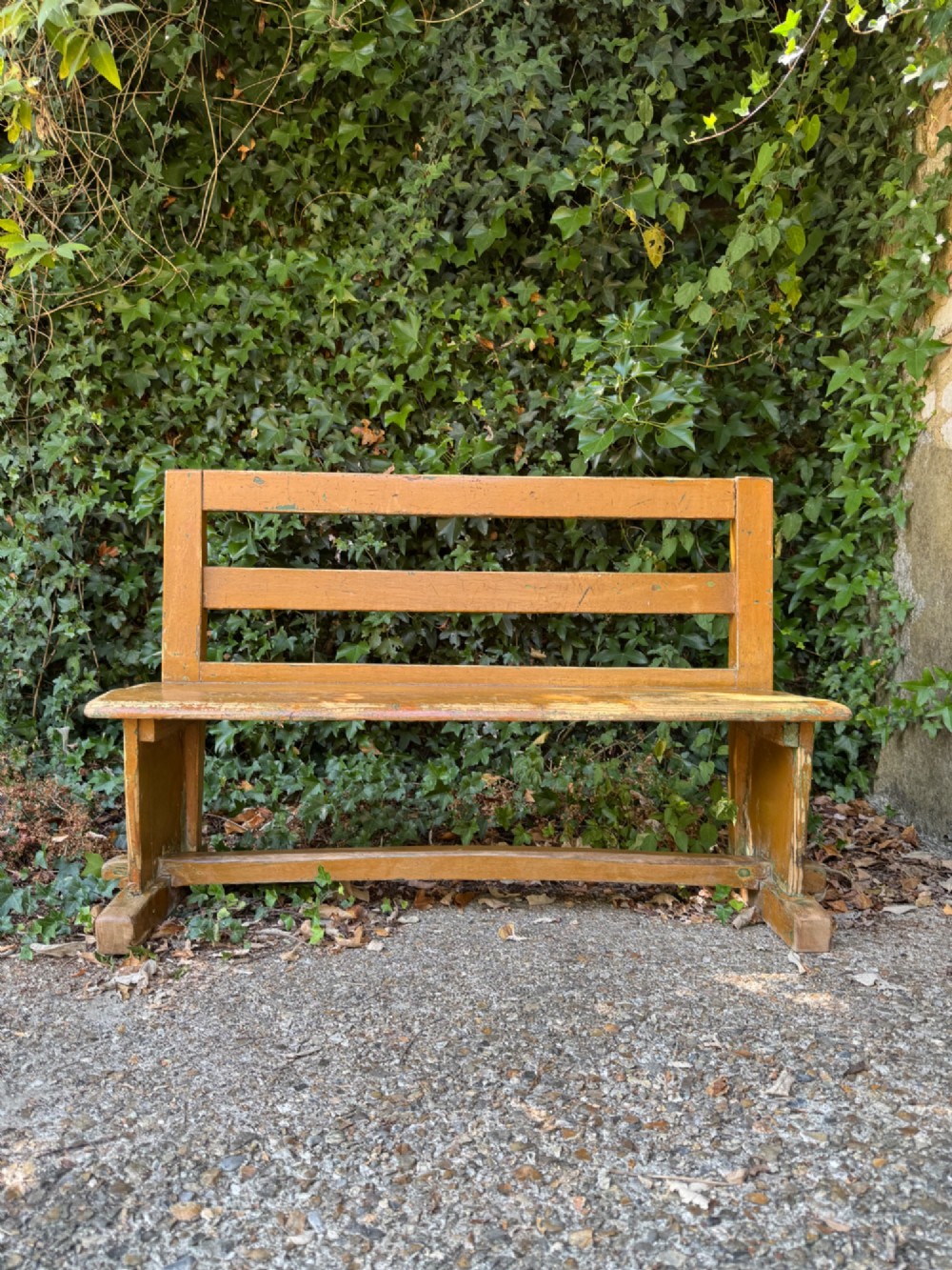 An antique rustic pine bench with a horizontal slat back, distressed mustard yellow painted finish, and solid end supports joined by a lower stretcher. View II