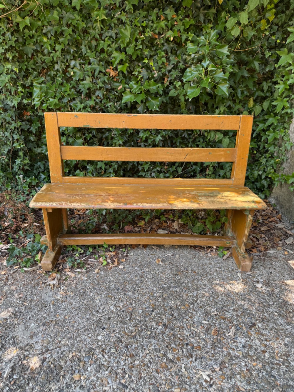 An antique rustic pine bench with a horizontal slat back, distressed mustard yellow painted finish, and solid end supports joined by a lower stretcher. View III