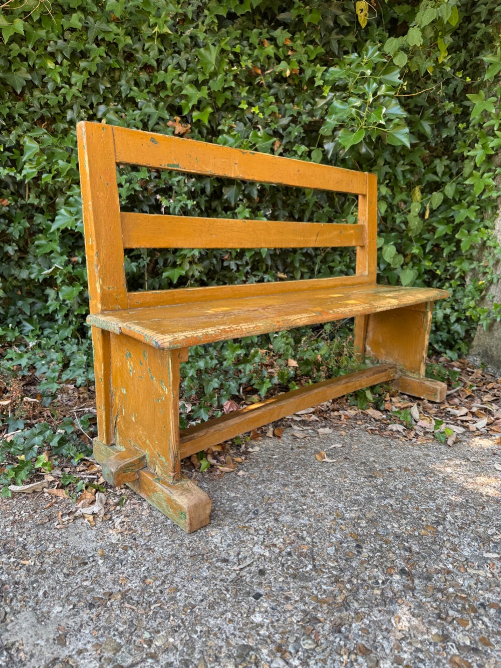 An antique rustic pine bench with a horizontal slat back, distressed mustard yellow painted finish, and solid end supports joined by a lower stretcher. View IV