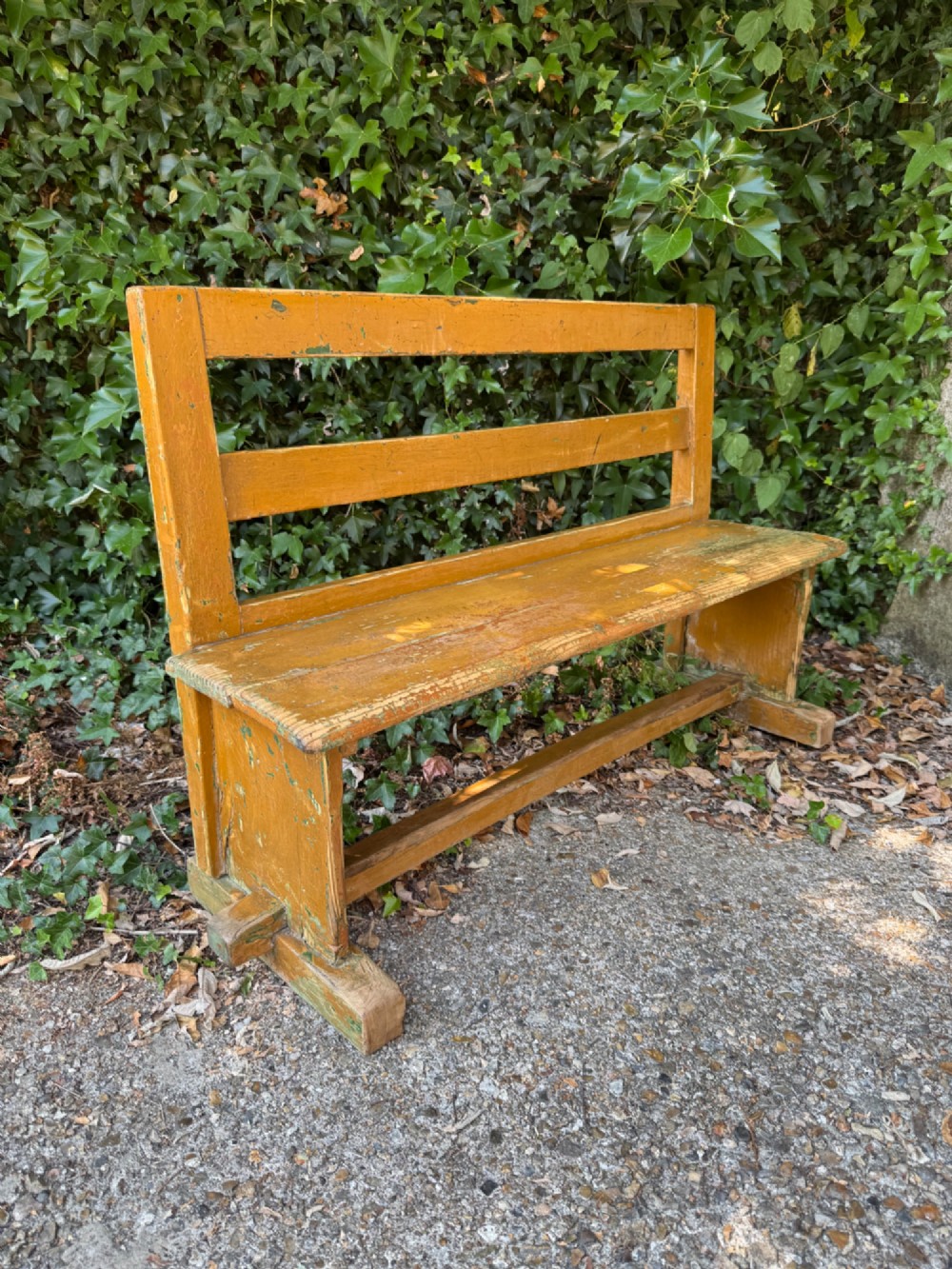 An antique rustic pine bench with a horizontal slat back, distressed mustard yellow painted finish, and solid end supports joined by a lower stretcher. View V