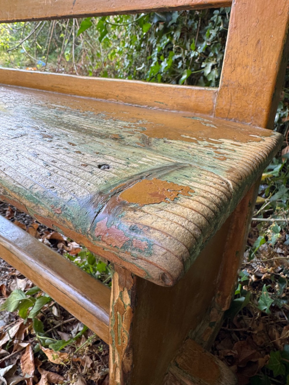 An antique rustic pine bench with a horizontal slat back, distressed mustard yellow painted finish, and solid end supports joined by a lower stretcher. View VI