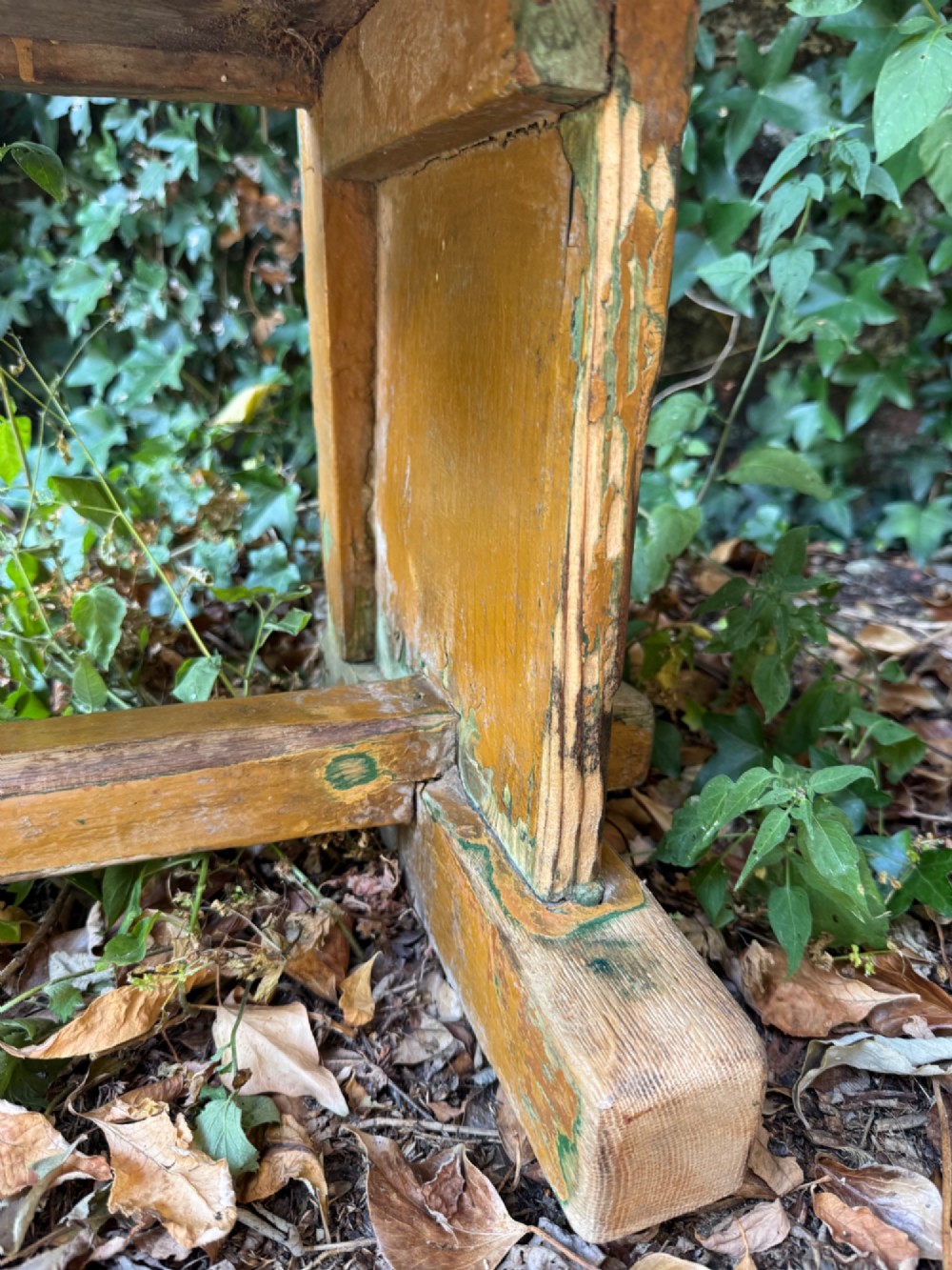 An antique rustic pine bench with a horizontal slat back, distressed mustard yellow painted finish, and solid end supports joined by a lower stretcher. View VII