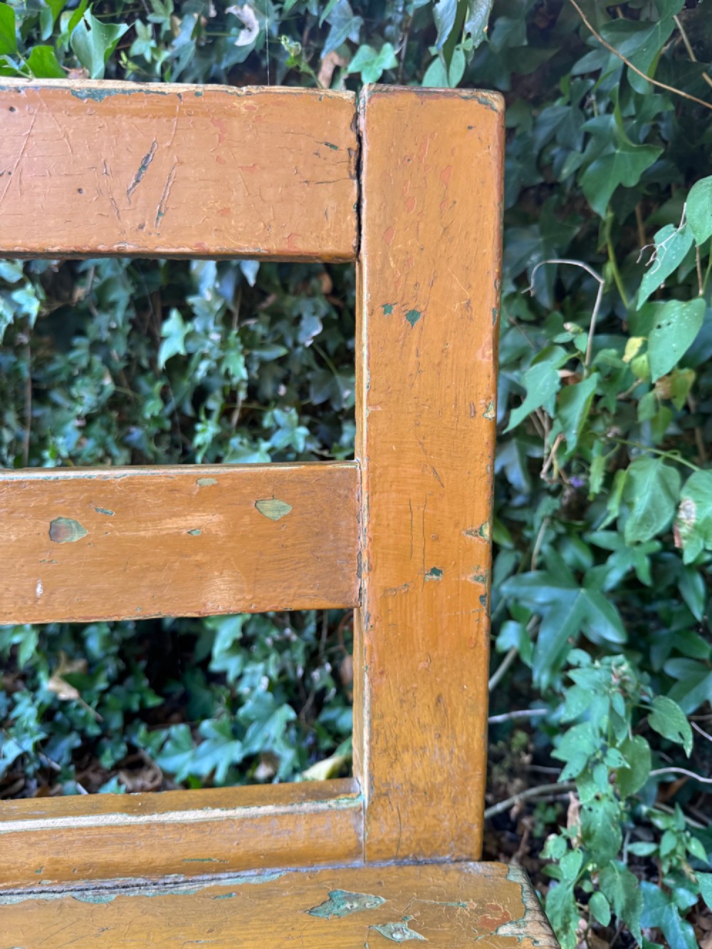 An antique rustic pine bench with a horizontal slat back, distressed mustard yellow painted finish, and solid end supports joined by a lower stretcher. View VIII