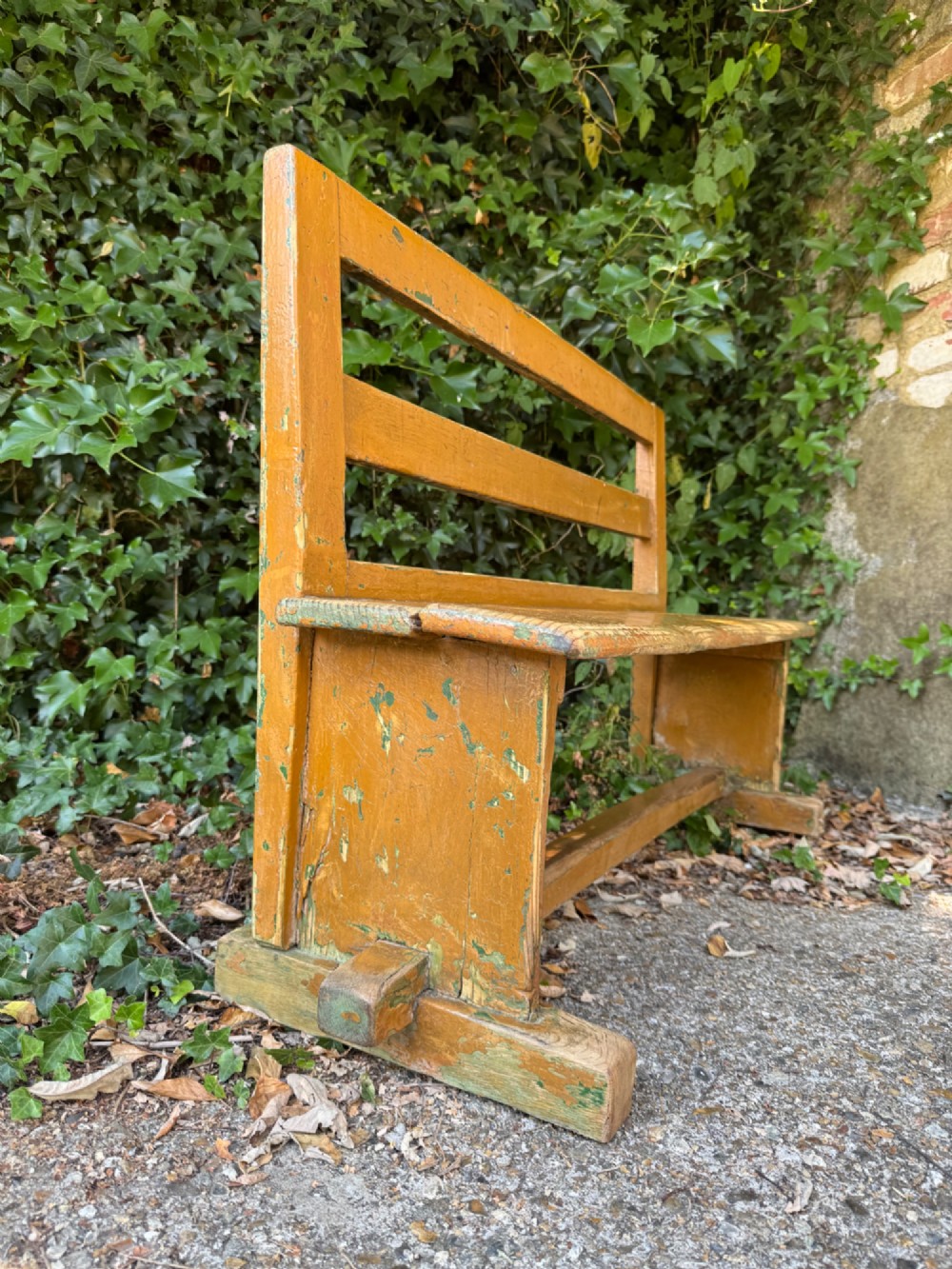 An antique rustic pine bench with a horizontal slat back, distressed mustard yellow painted finish, and solid end supports joined by a lower stretcher. View IX