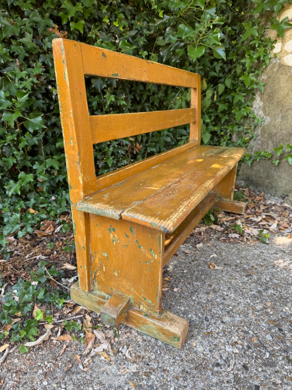 An antique rustic pine bench with a horizontal slat back, distressed mustard yellow painted finish, and solid end supports joined by a lower stretcher. View X