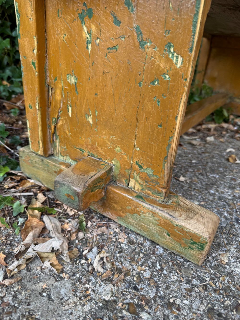 An antique rustic pine bench with a horizontal slat back, distressed mustard yellow painted finish, and solid end supports joined by a lower stretcher. View XII
