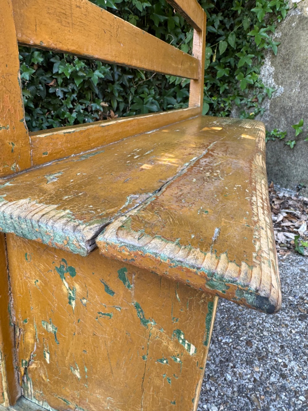 An antique rustic pine bench with a horizontal slat back, distressed mustard yellow painted finish, and solid end supports joined by a lower stretcher. View XIV