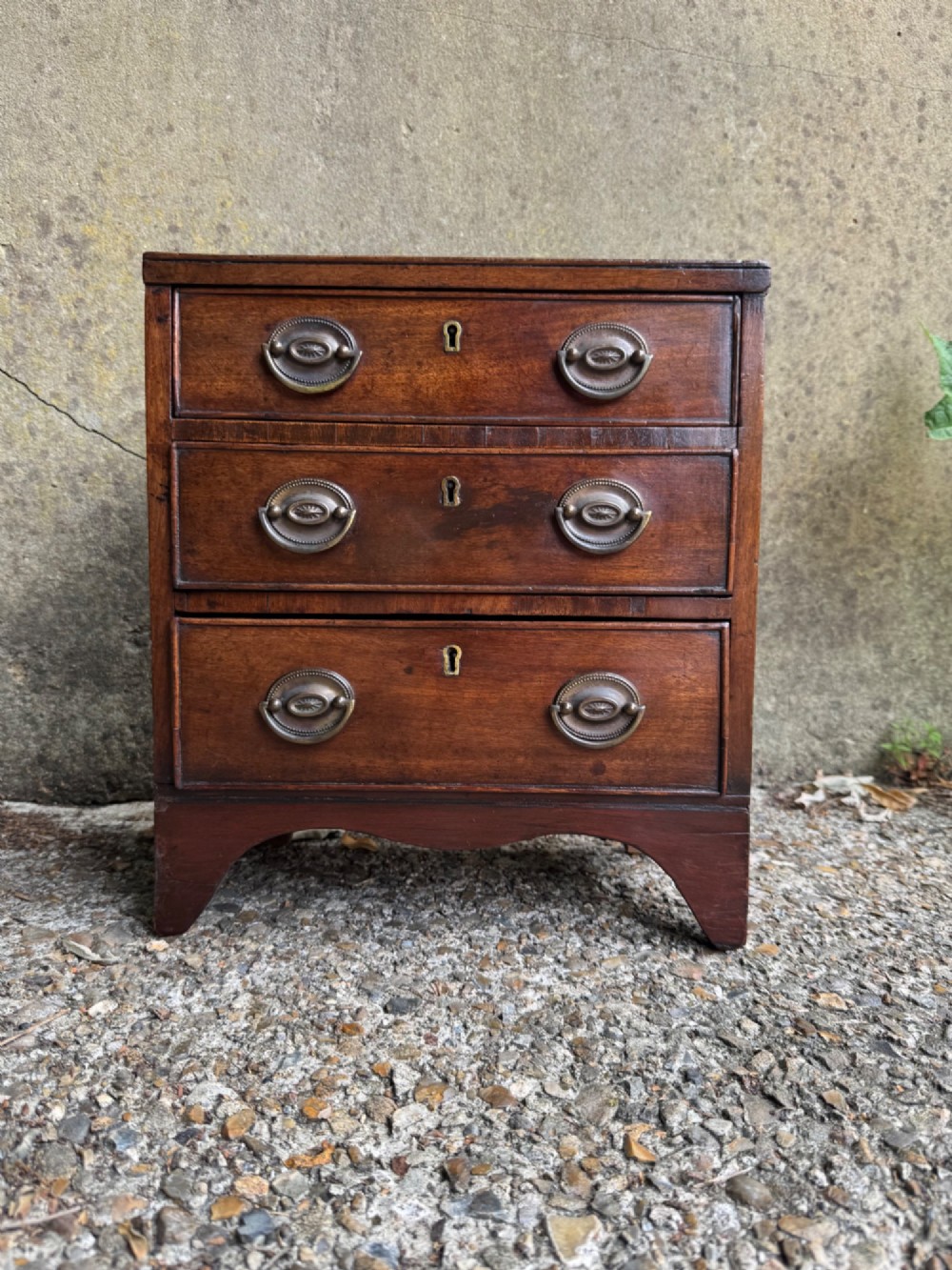 A miniature antique mahogany apprentice chest featuring three drawers with brass oval drop handles and keyhole escutcheons resting on a shaped apron base.