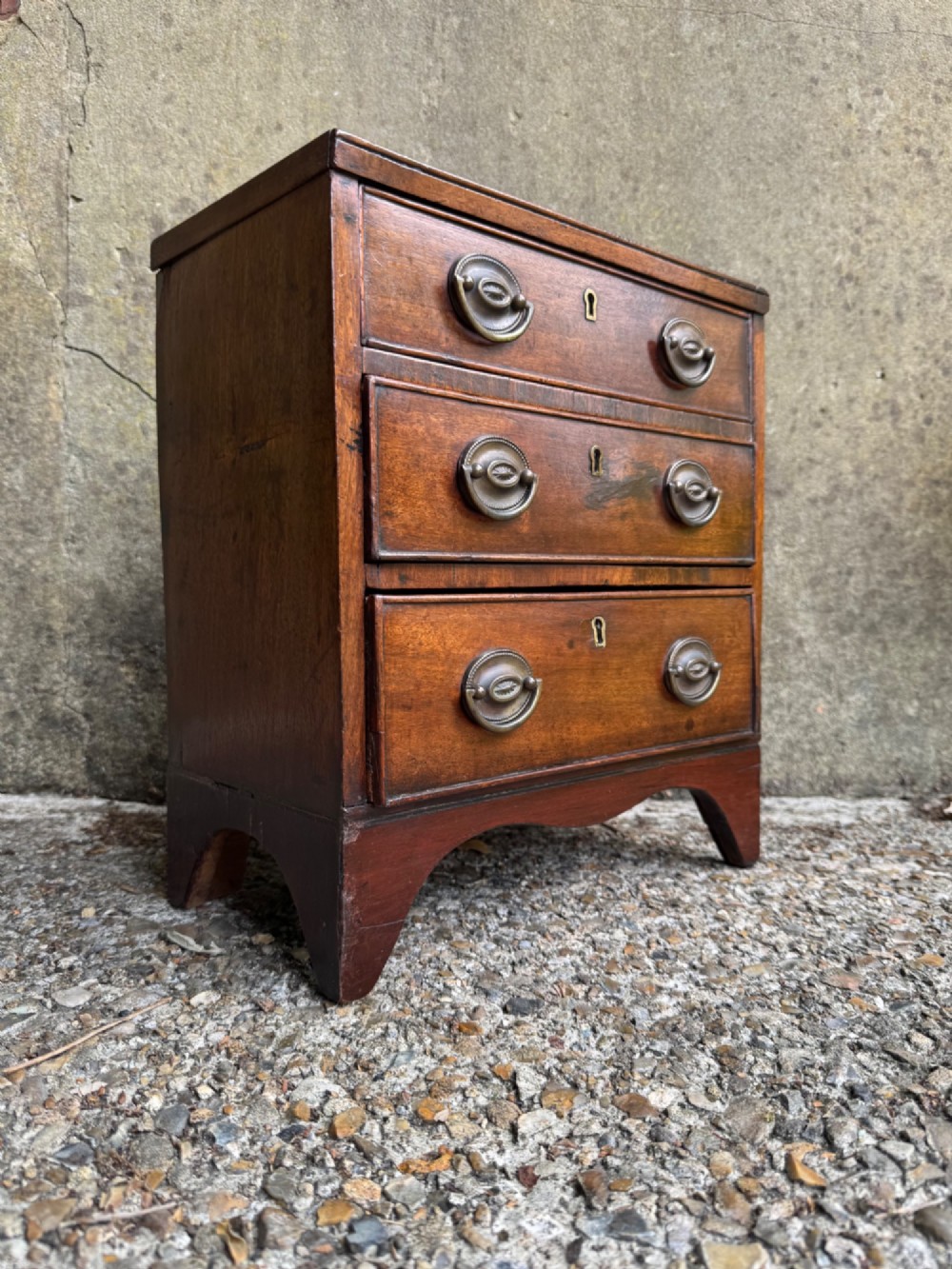 A miniature antique mahogany apprentice chest featuring three drawers with brass oval drop handles and keyhole escutcheons resting on a shaped apron base. View II