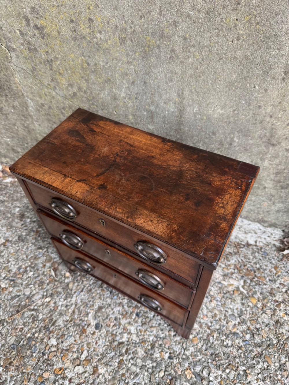 A miniature antique mahogany apprentice chest featuring three drawers with brass oval drop handles and keyhole escutcheons resting on a shaped apron base. View III