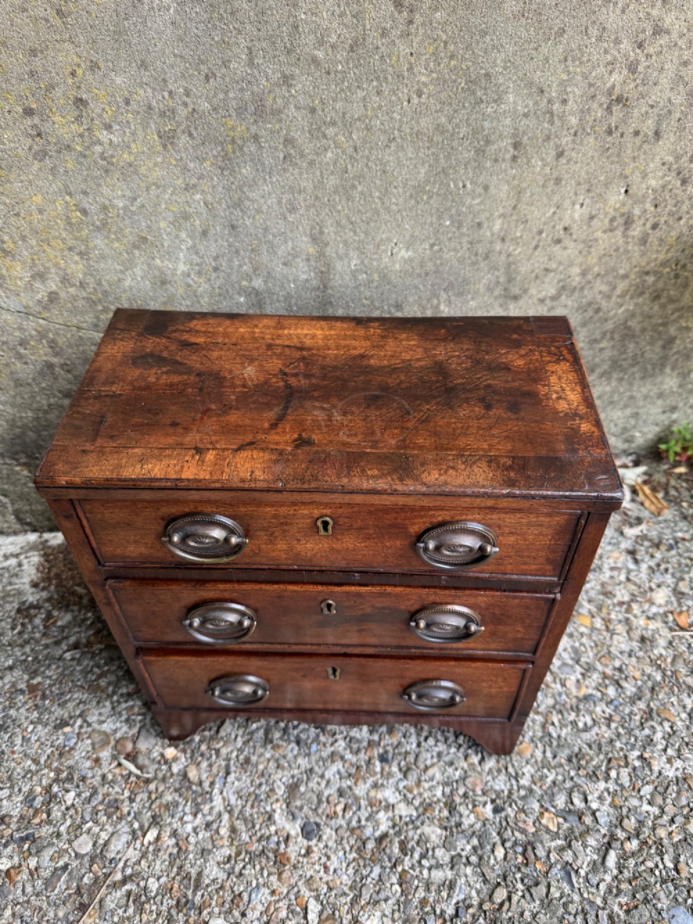 A miniature antique mahogany apprentice chest featuring three drawers with brass oval drop handles and keyhole escutcheons resting on a shaped apron base. View IV