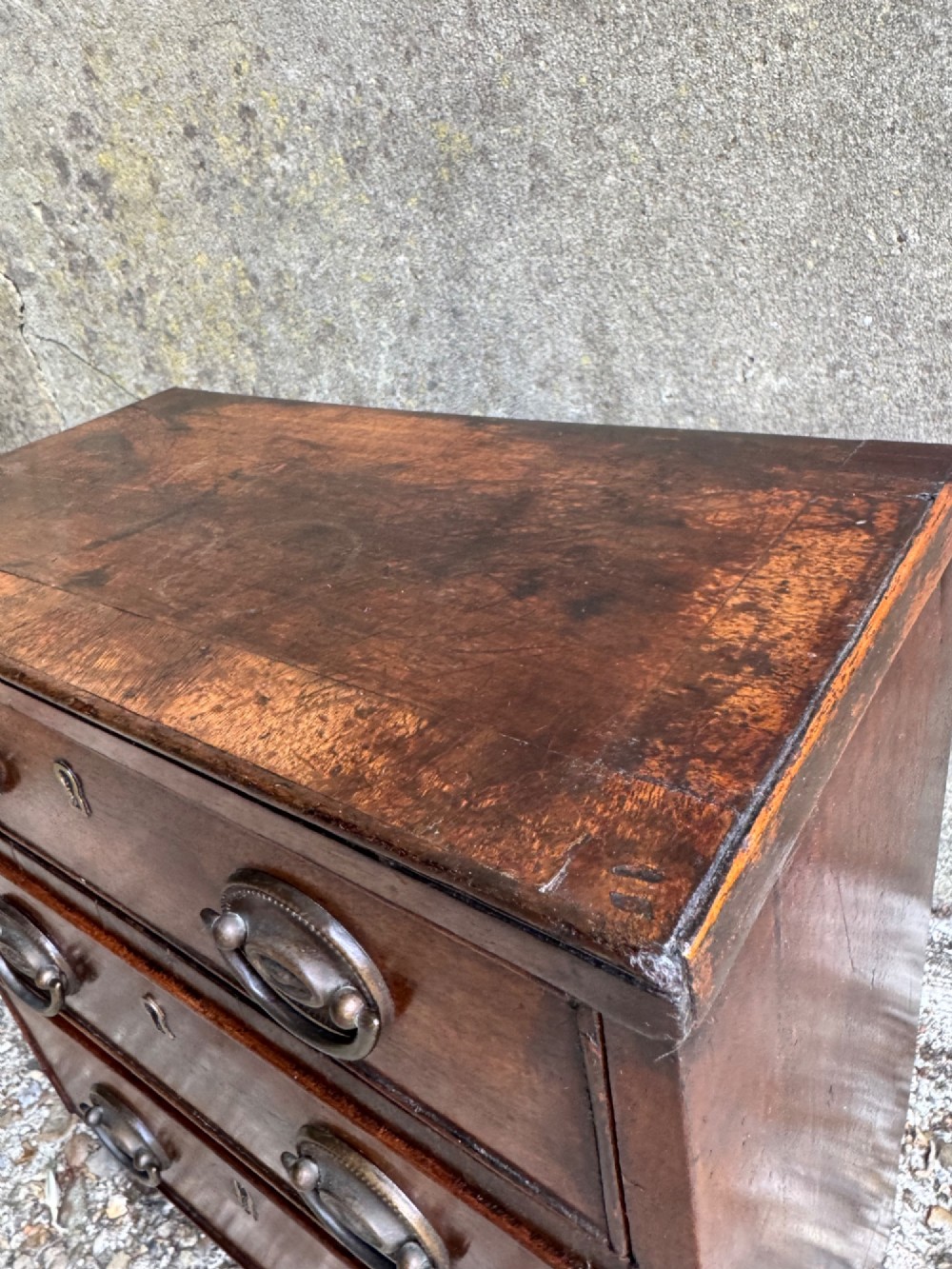 A miniature antique mahogany apprentice chest featuring three drawers with brass oval drop handles and keyhole escutcheons resting on a shaped apron base. View VI