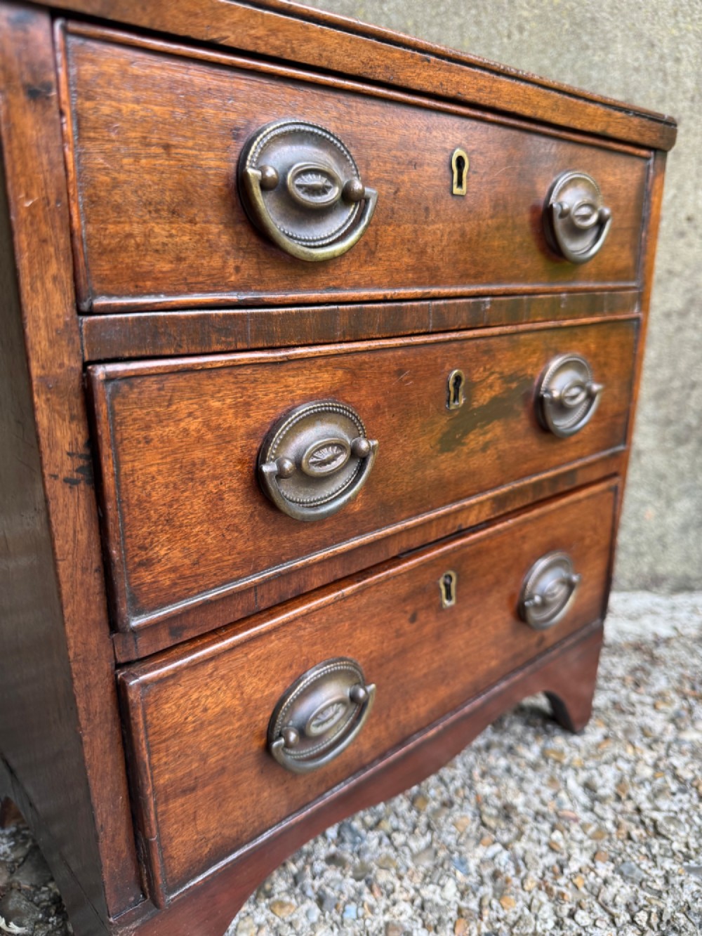 A miniature antique mahogany apprentice chest featuring three drawers with brass oval drop handles and keyhole escutcheons resting on a shaped apron base. View VII
