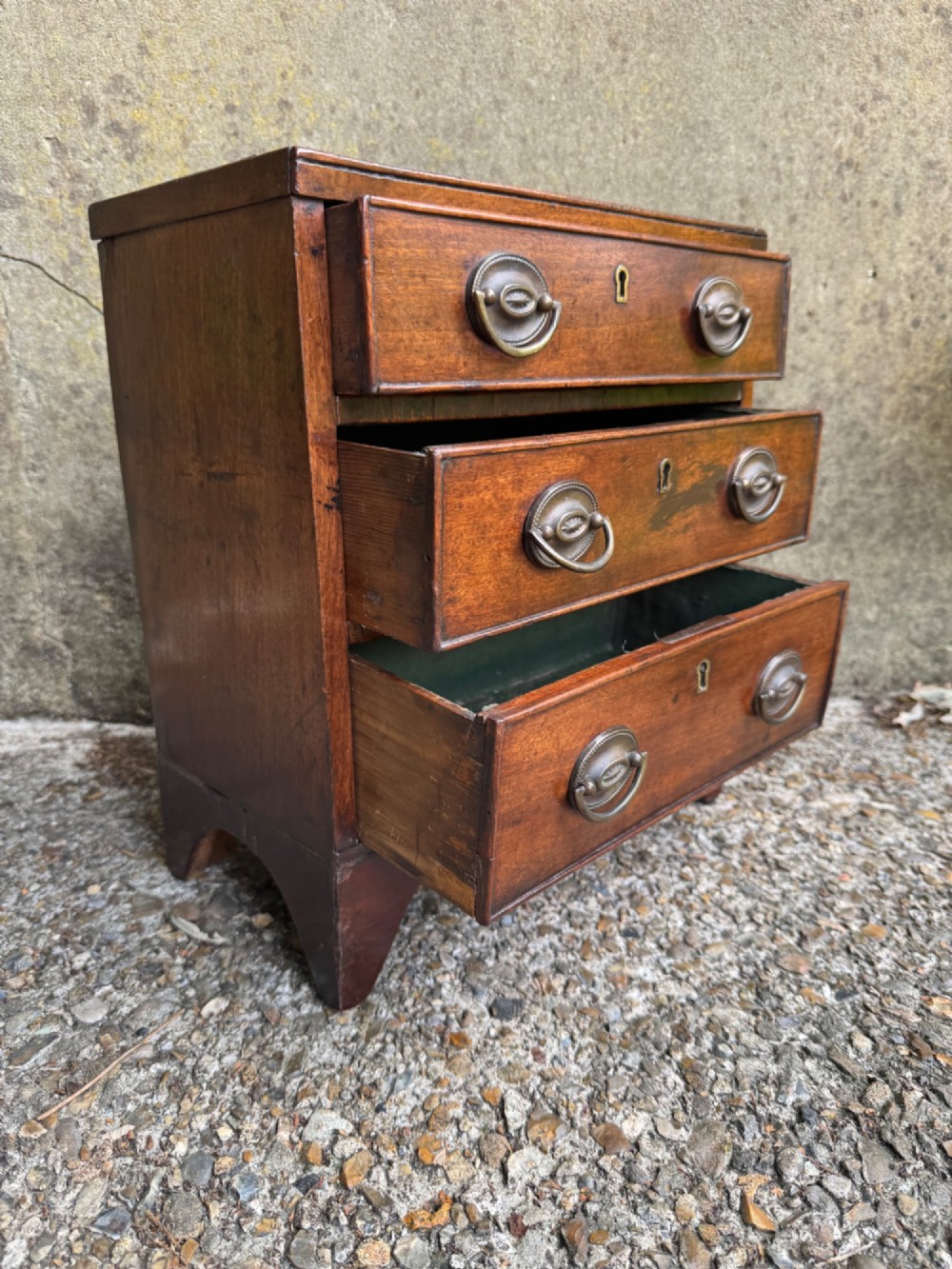 A miniature antique mahogany apprentice chest featuring three drawers with brass oval drop handles and keyhole escutcheons resting on a shaped apron base. View VIII