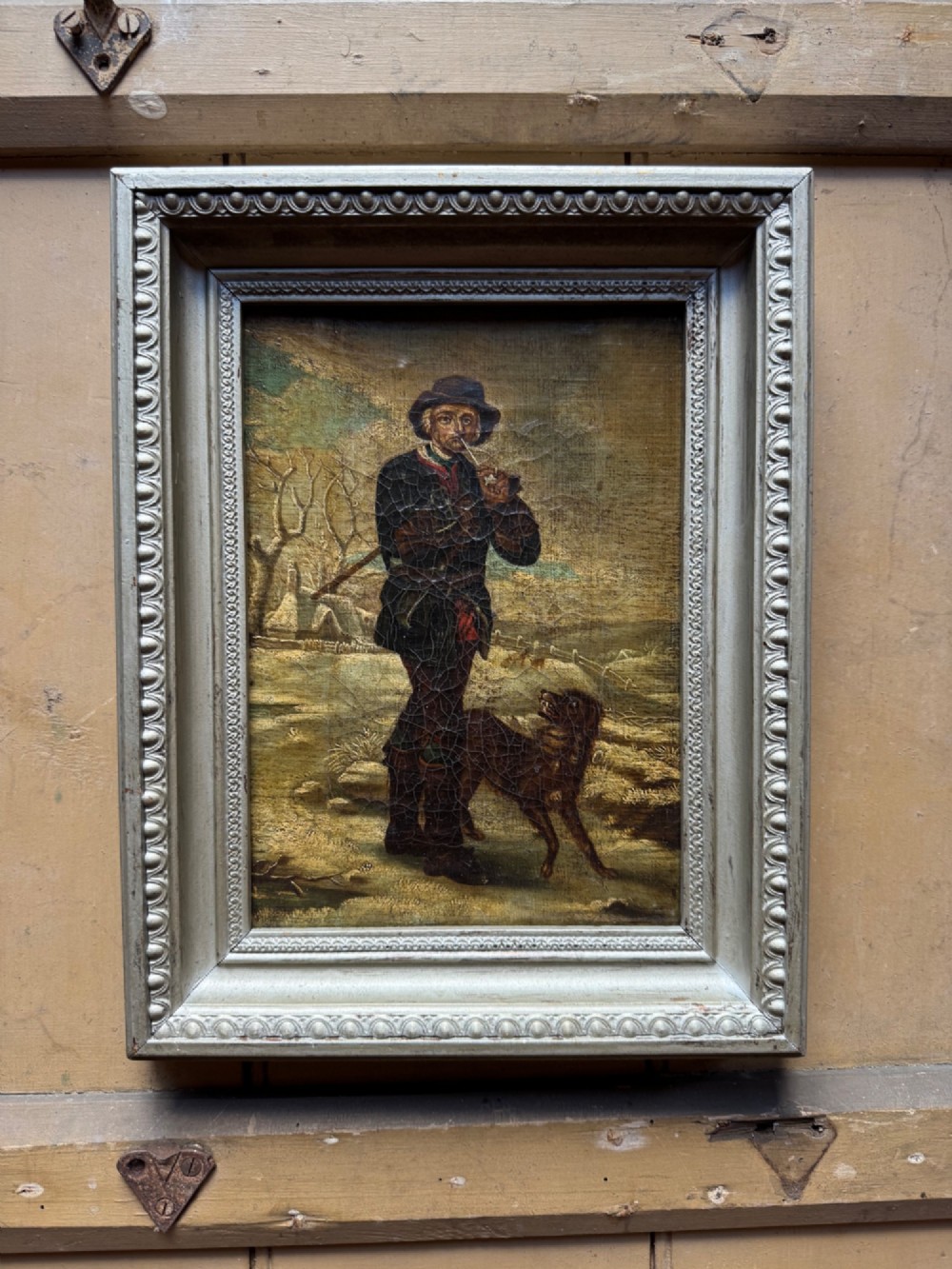 An antique oil painting depicting a standing man with a pipe and his dog in a countryside landscape, housed in a deep silvered moulded frame.