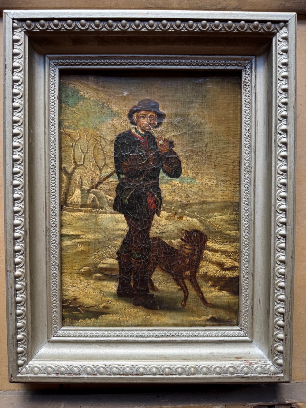 An antique oil painting depicting a standing man with a pipe and his dog in a countryside landscape, housed in a deep silvered moulded frame. View II