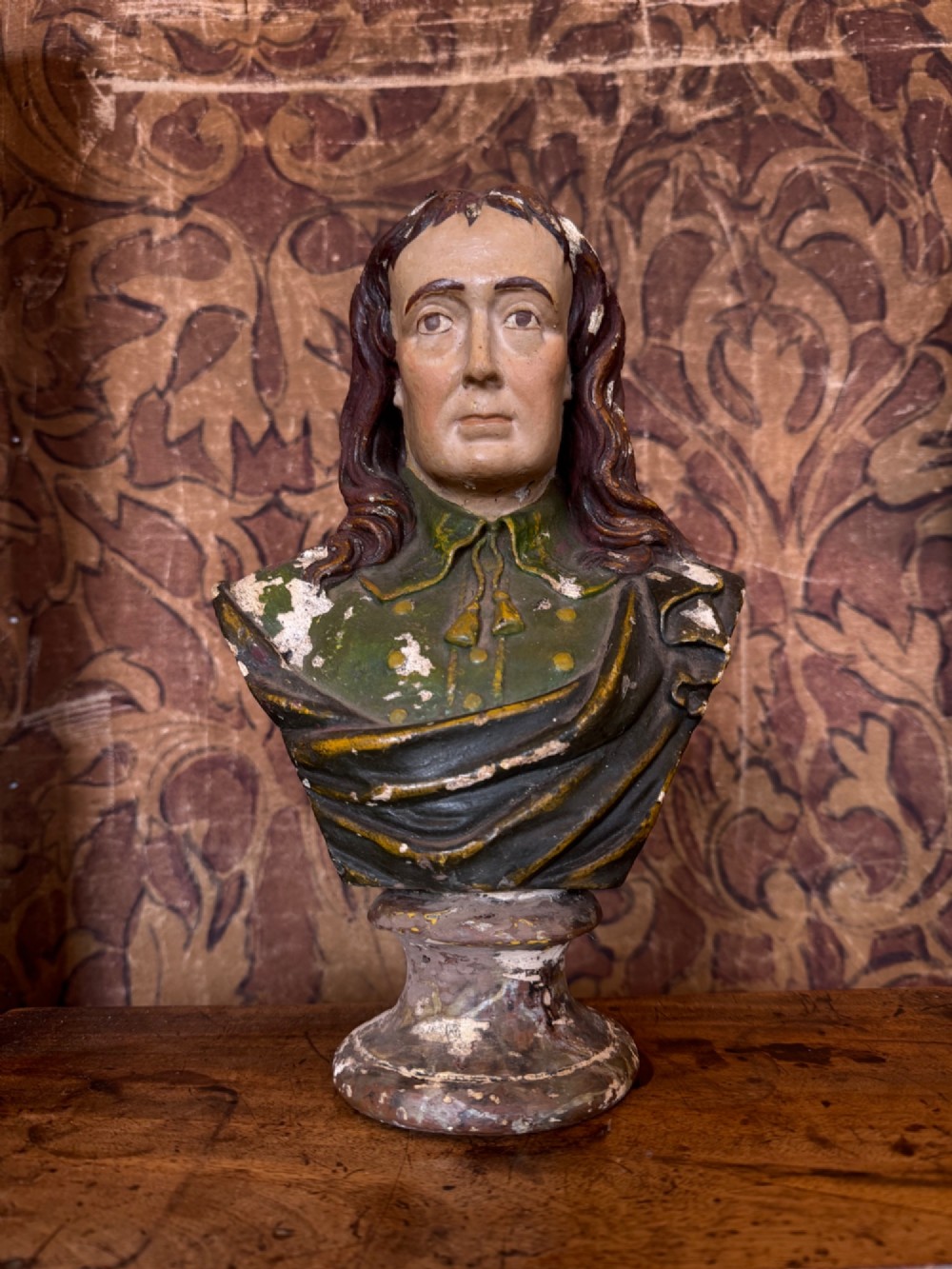 A polychrome painted bust depicting John Milton with long brown hair, a collared shirt with tassels, and a grey socle pedestal base.