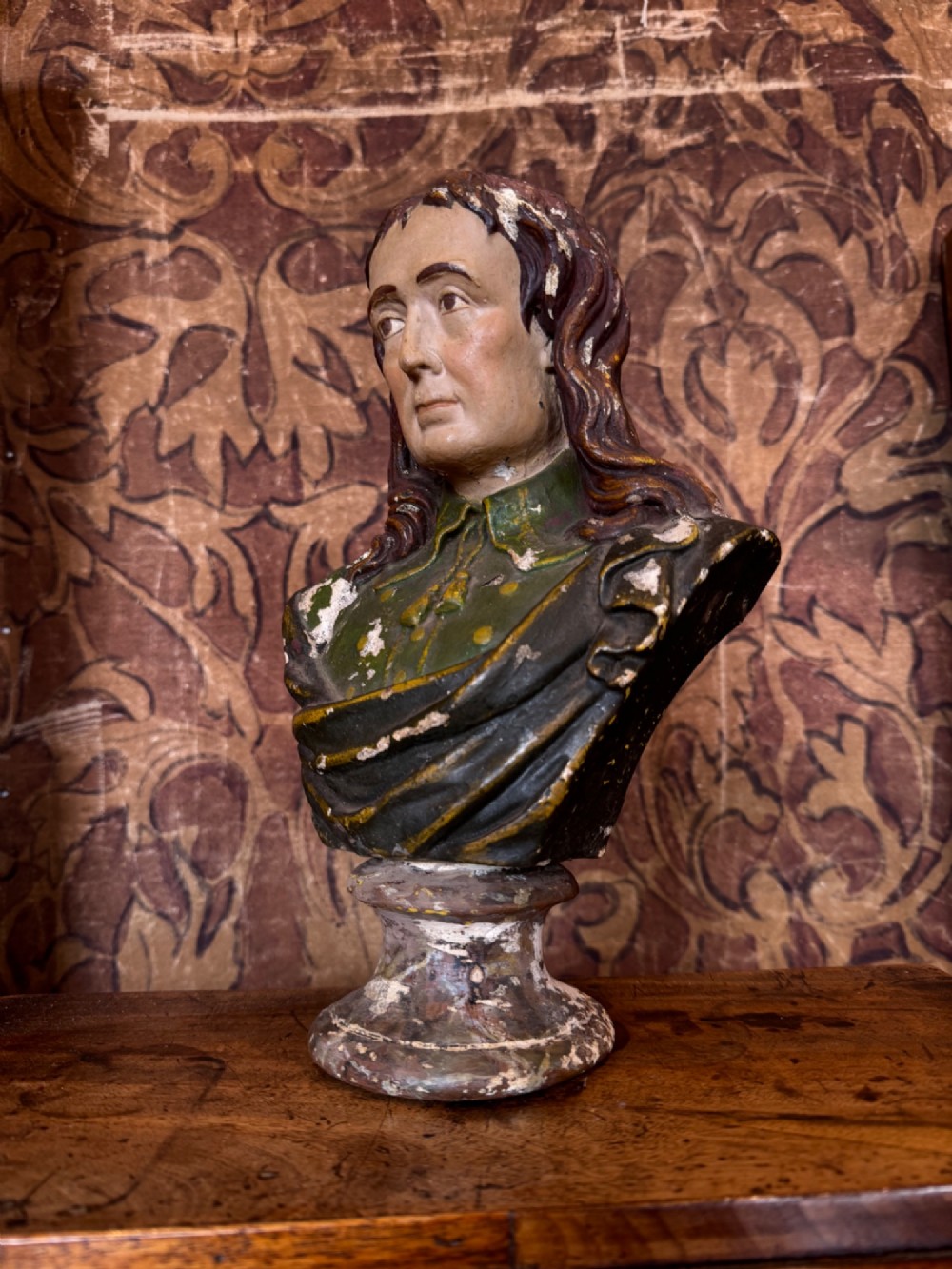 A polychrome painted bust depicting John Milton with long brown hair, a collared shirt with tassels, and a grey socle pedestal base. View II
