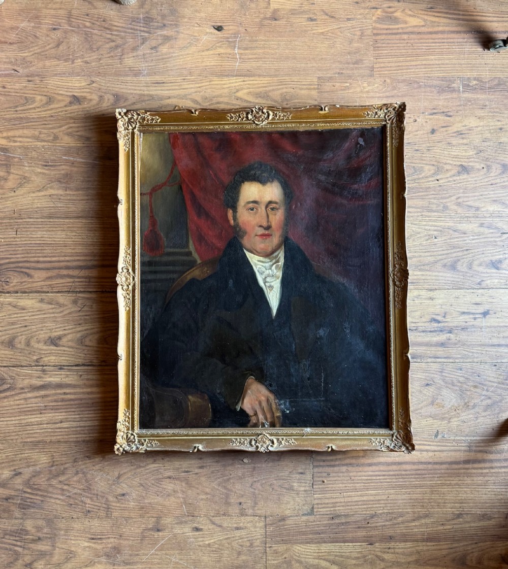 An antique oil painting depicting a gentleman in a dark coat and white neckcloth seated before a red drape, housed in a moulded giltwood frame.
