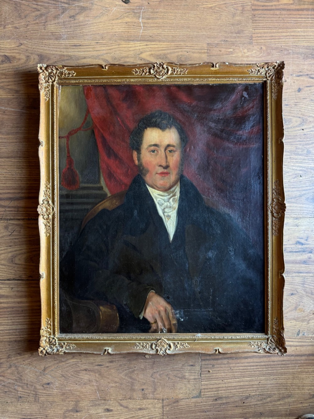 An antique oil painting depicting a gentleman in a dark coat and white neckcloth seated before a red drape, housed in a moulded giltwood frame. View II