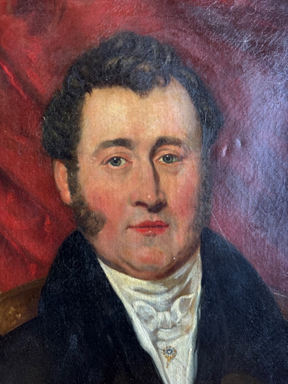An antique oil painting depicting a gentleman in a dark coat and white neckcloth seated before a red drape, housed in a moulded giltwood frame. View III