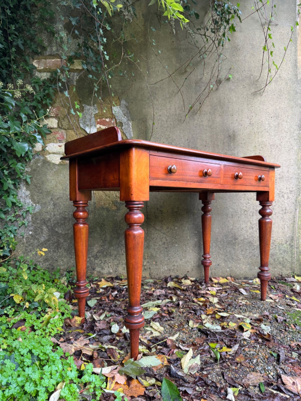An antique reddish-brown mahogany writing table featuring two drawers with wooden knob handles, turned legs, and a three-sided raised rear gallery.