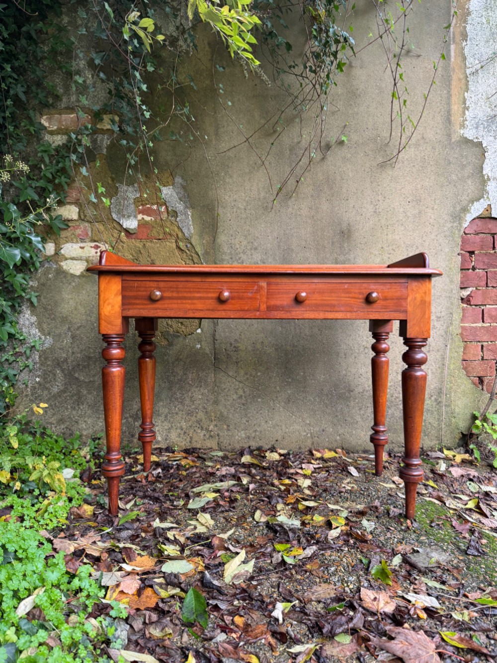 An antique reddish-brown mahogany writing table featuring two drawers with wooden knob handles, turned legs, and a three-sided raised rear gallery. View II
