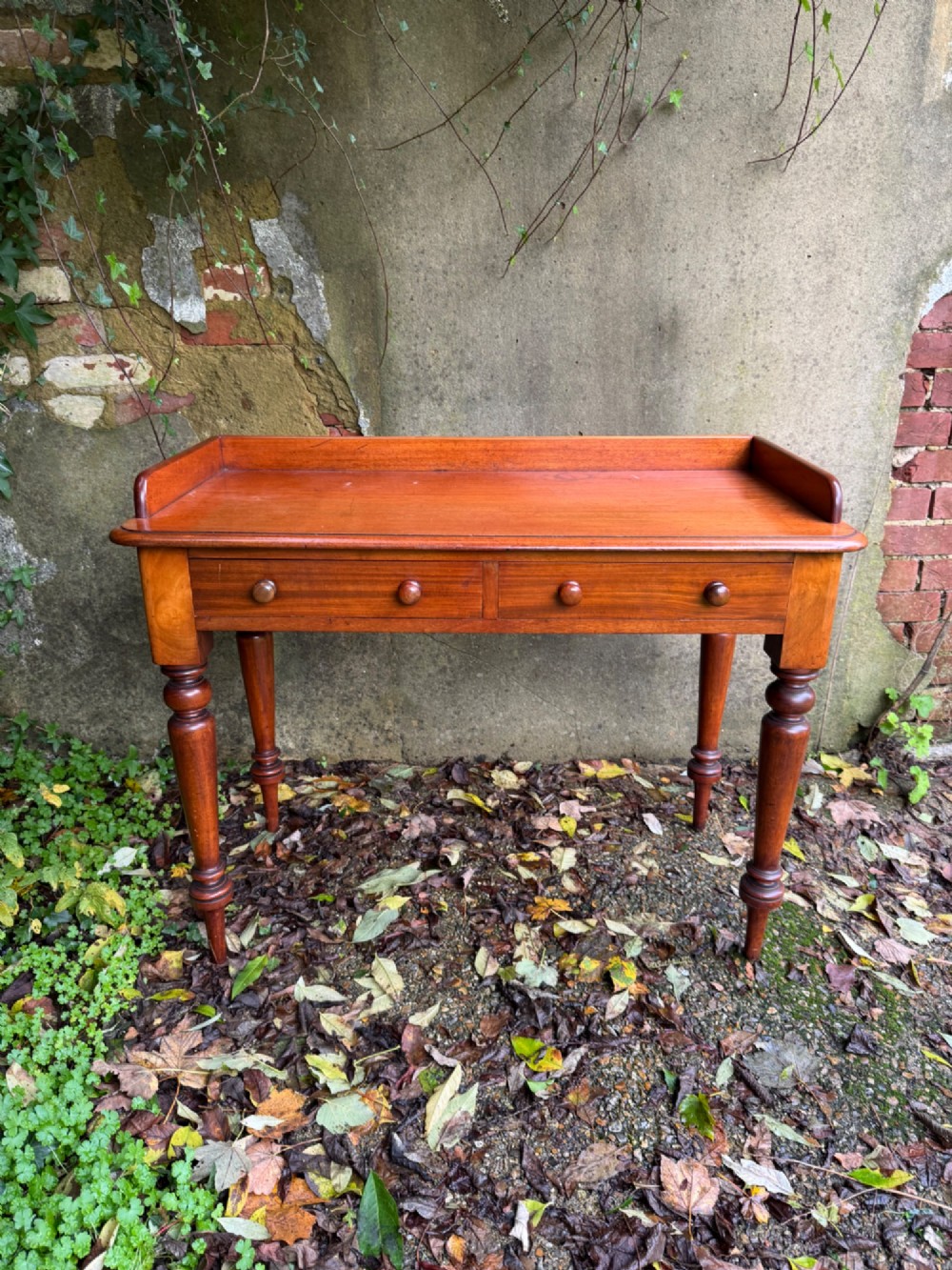 An antique reddish-brown mahogany writing table featuring two drawers with wooden knob handles, turned legs, and a three-sided raised rear gallery. View IV