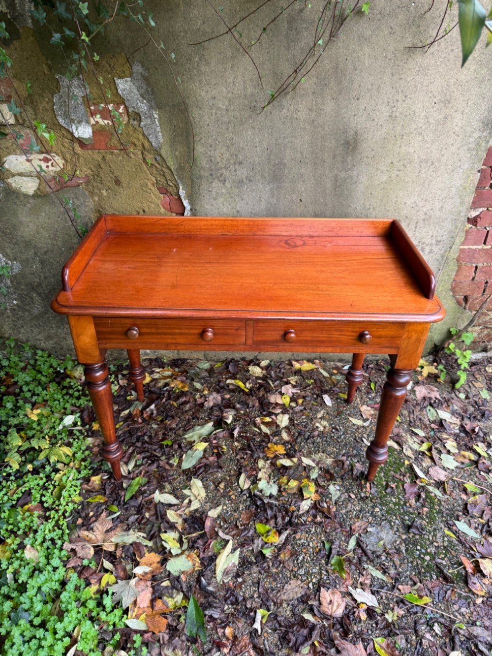 An antique reddish-brown mahogany writing table featuring two drawers with wooden knob handles, turned legs, and a three-sided raised rear gallery. View VII