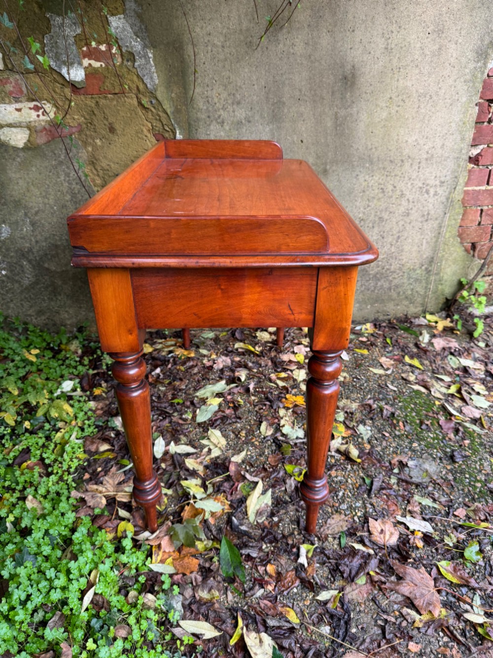An antique reddish-brown mahogany writing table featuring two drawers with wooden knob handles, turned legs, and a three-sided raised rear gallery. View XI
