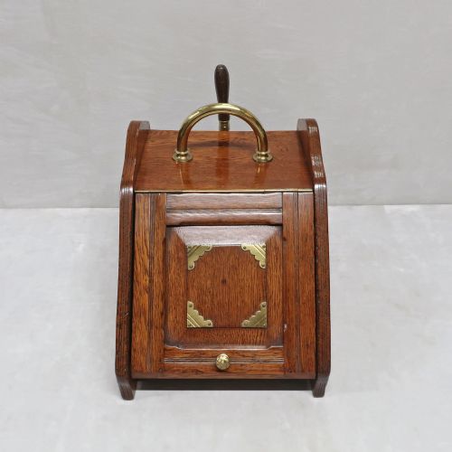 fig 1. A Victorian oak coal scuttle featuring a slanted drop-down front door decorated with brass corner mounts and a brass carrying handle on top.