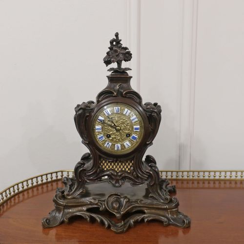 fig 1. A patinated dark bronze French Rococo style mantel clock featuring scrolling acanthus motifs, a floral finial, and blue Roman numerals on individual enamel cartouches.