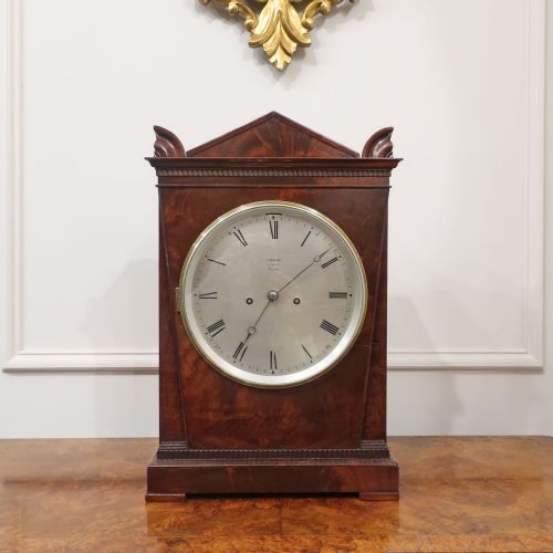 fig 1. A Georgian flame mahogany bracket clock featuring a circular white dial with Roman numerals, a pointed architectural pediment, and brass side frets.