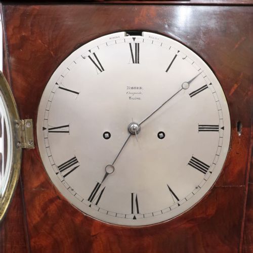 fig 2. A Georgian flame mahogany bracket clock featuring a circular white dial with Roman numerals, a pointed architectural pediment, and brass side frets.