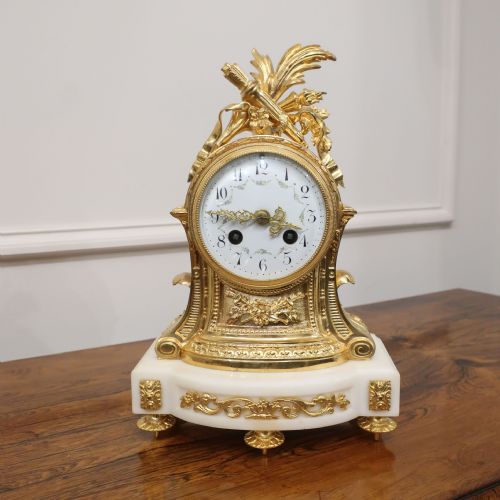 fig 1. An ornate French gilt bronze and white marble mantel clock featuring a white enamel dial topped with leaf motifs.