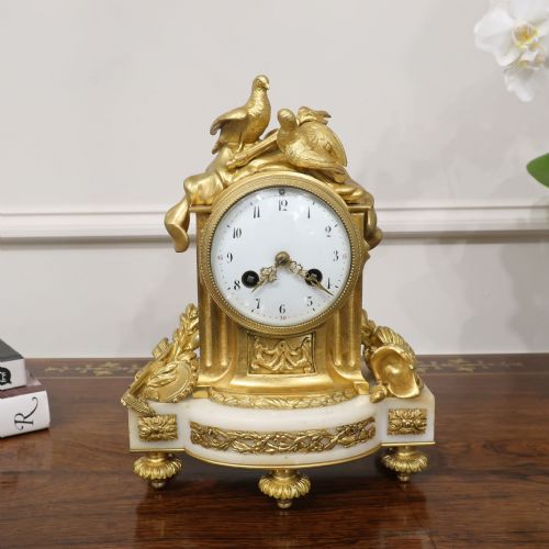 fig 1. A French Louis XVI style gilt bronze and white marble mantel clock featuring a pair of sculpted doves atop a circular enamel dial flanked by fluted pillars.