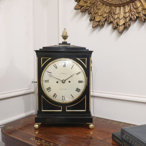 fig 1. An ebonised William IV bracket clock featuring brass inlays, a pineapple finial, lion mask side handles, ball feet, and a white circular dial with Roman numerals signed J. Bull Bedford.