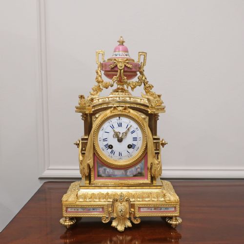 fig 1. A French gilt bronze mantel clock featuring an urn top finial, pink hand-painted porcelain panels, and a white enamel dial with Roman numerals.