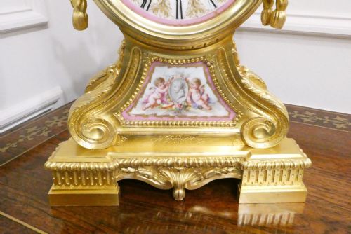 fig 5. A French Louis XV style gilt bronze mantel clock featuring a hand-painted porcelain dial, lower cherub panel, scroll feet, and an ornamental urn finial.