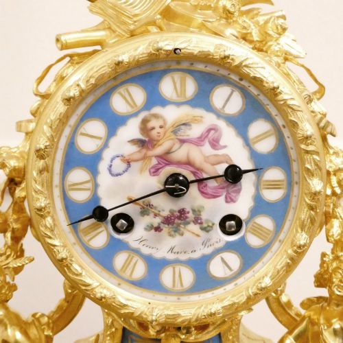 fig 1. An ornate antique French gilt bronze mantel clock featuring decorative cherub figures, a recumbent goat sculpture, and blue porcelain panels painted with cherubs and floral motifs.