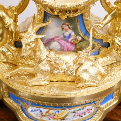 fig 2. An ornate antique French gilt bronze mantel clock featuring decorative cherub figures, a recumbent goat sculpture, and blue porcelain panels painted with cherubs and floral motifs.