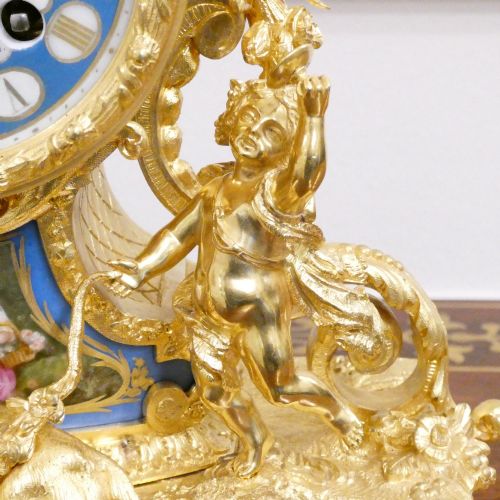fig 3. An ornate antique French gilt bronze mantel clock featuring decorative cherub figures, a recumbent goat sculpture, and blue porcelain panels painted with cherubs and floral motifs.