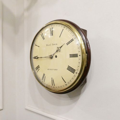 fig 1. A circular Georgian wall clock featuring a cream dial with black Roman numerals, blued steel hands, a polished brass bezel, and a dark mahogany wood surround.
