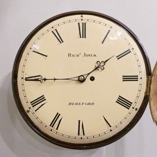 fig 2. A circular Georgian wall clock featuring a cream dial with black Roman numerals, blued steel hands, a polished brass bezel, and a dark mahogany wood surround.