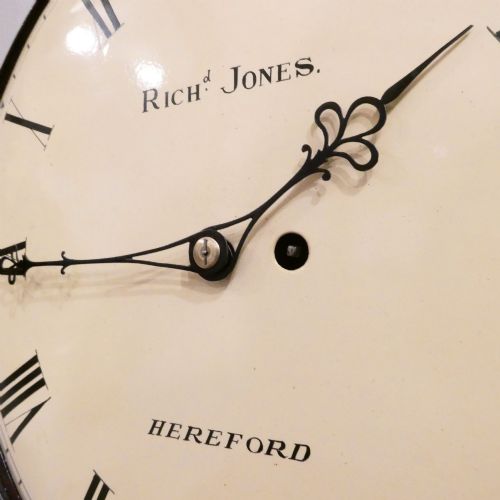fig 3. A circular Georgian wall clock featuring a cream dial with black Roman numerals, blued steel hands, a polished brass bezel, and a dark mahogany wood surround.