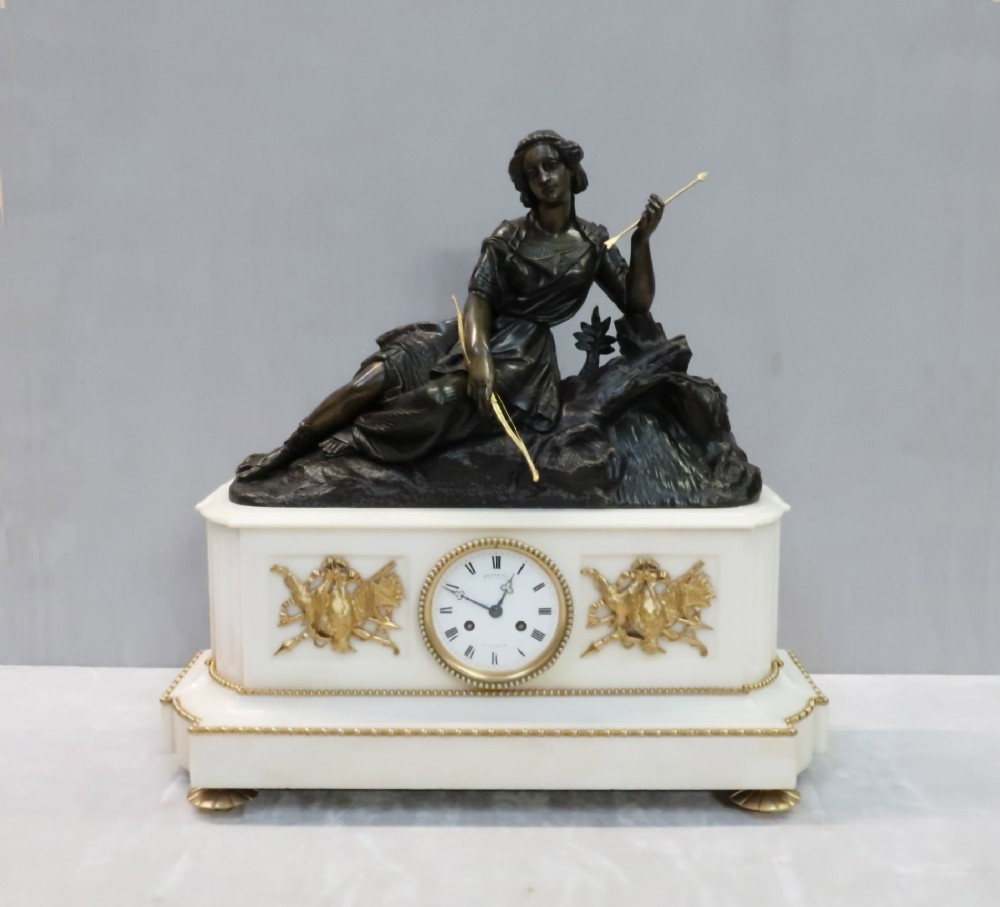 An antique French Napoleon III mantel clock featuring a dark patinated bronze figure of Diana the Huntress holding a bow, set atop a rectangular white marble base with gilt-bronze mounts.