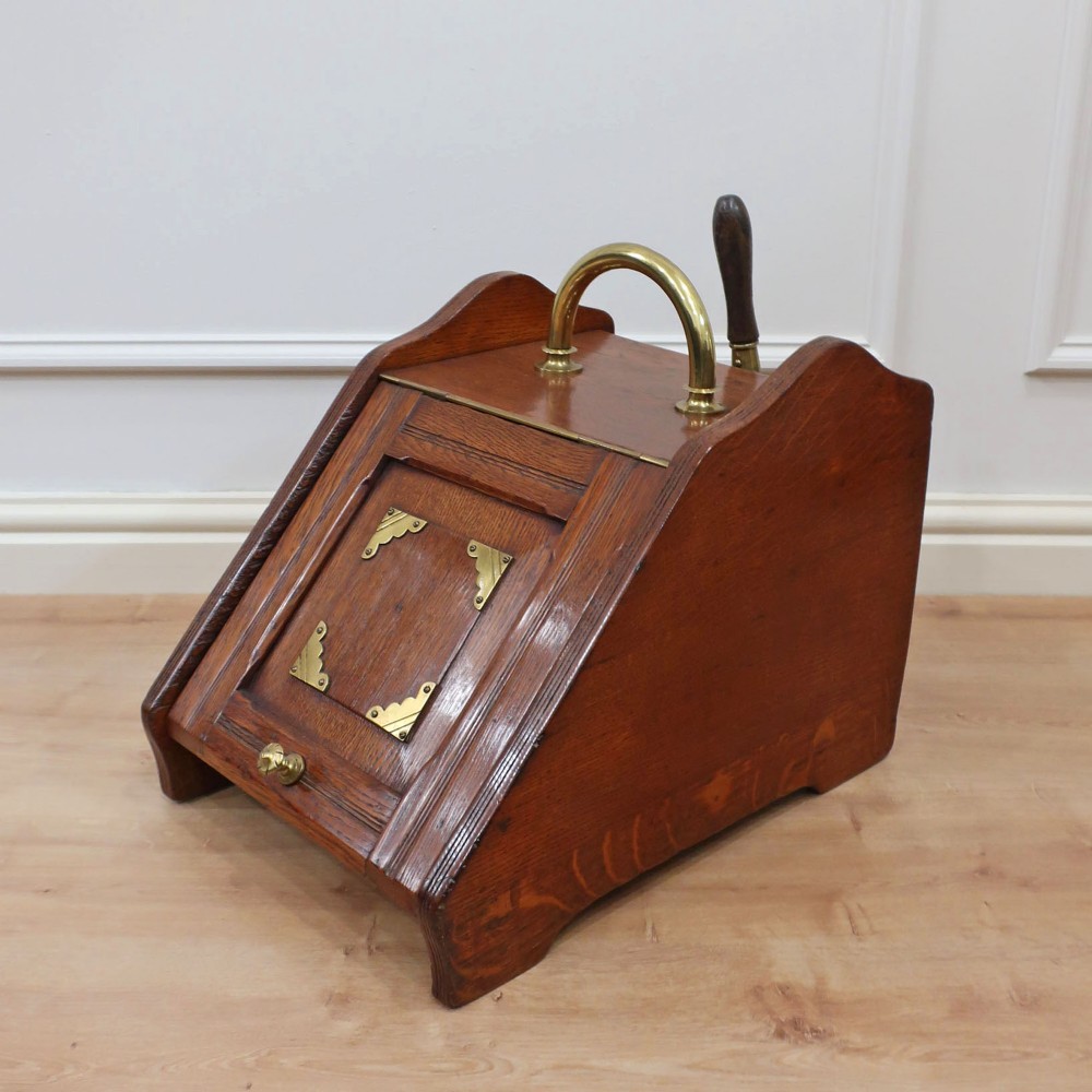 A Victorian oak coal scuttle featuring a slanted drop-down front door decorated with brass corner mounts and a brass carrying handle on top.